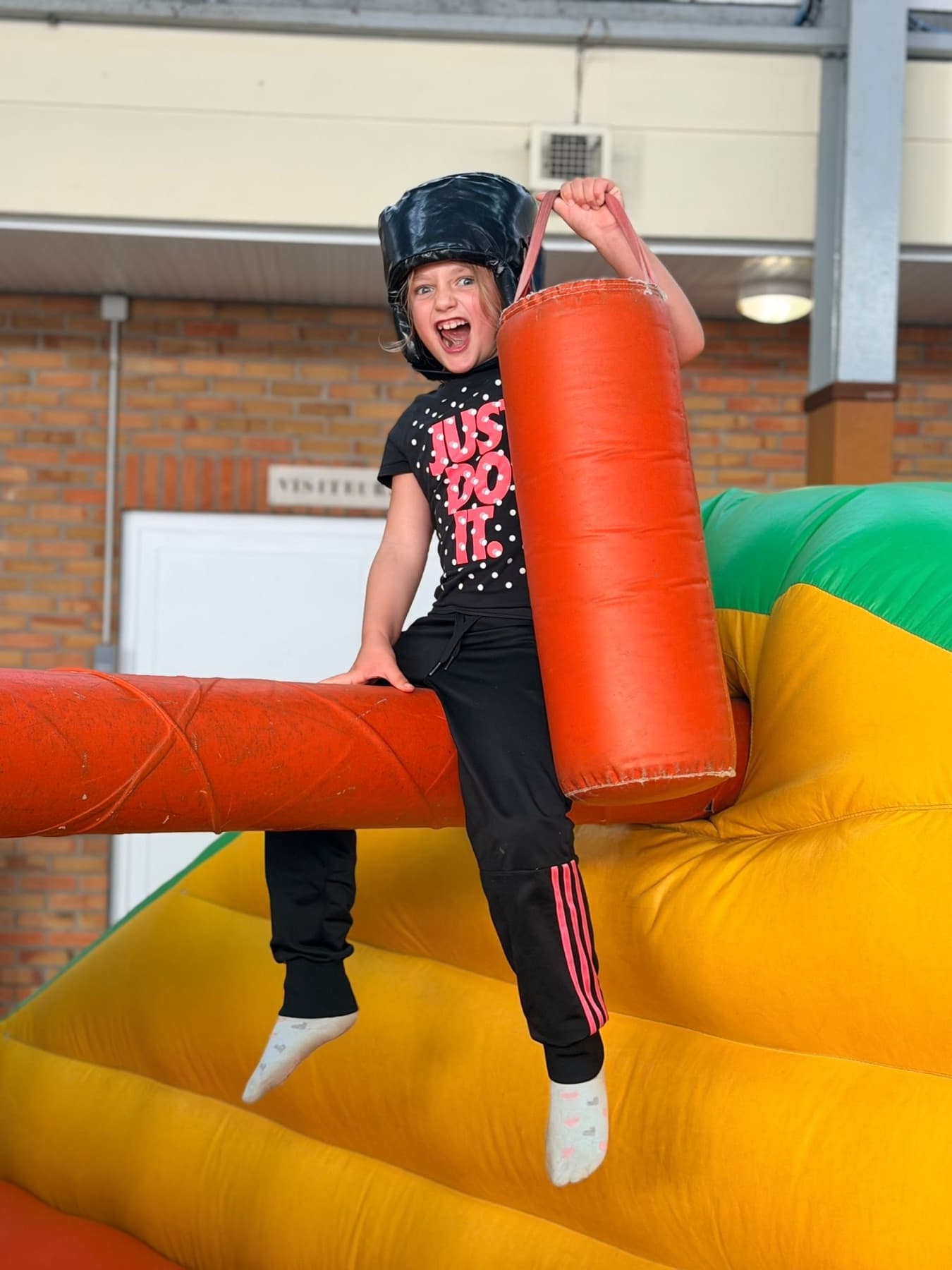 Enfant souriante avec un casque sur un parcours gonflable de gladiateurs.